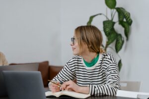 An adult student writing in a notebook beside a laptop at a desk