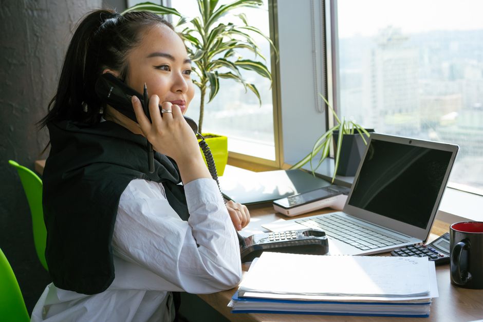 A woman making a phone call at a desk with papers and a laptop